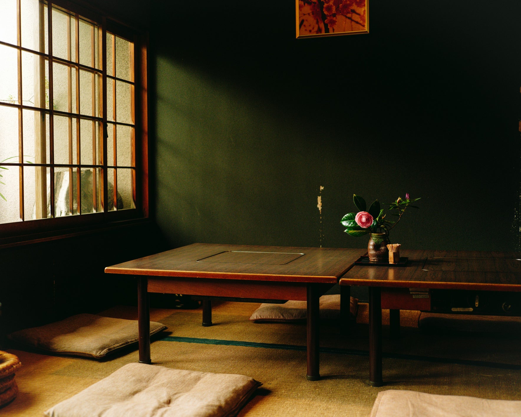 A peaceful, minimalist Japanese tea room, featuring a low wooden table, seat cushions, and a simple floral arrangement in a vase by the window, with sunlight streaming in softly.