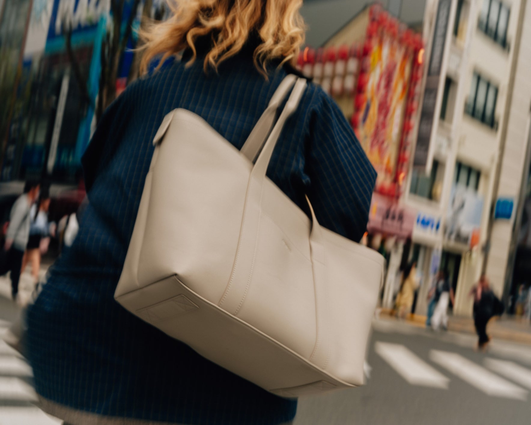 A close-up shot of a woman carrying a metro tote in ivory (vegan leather) over her shoulder as she walks through a busy Japanese street, with colourful buildings and people in the blurred background.