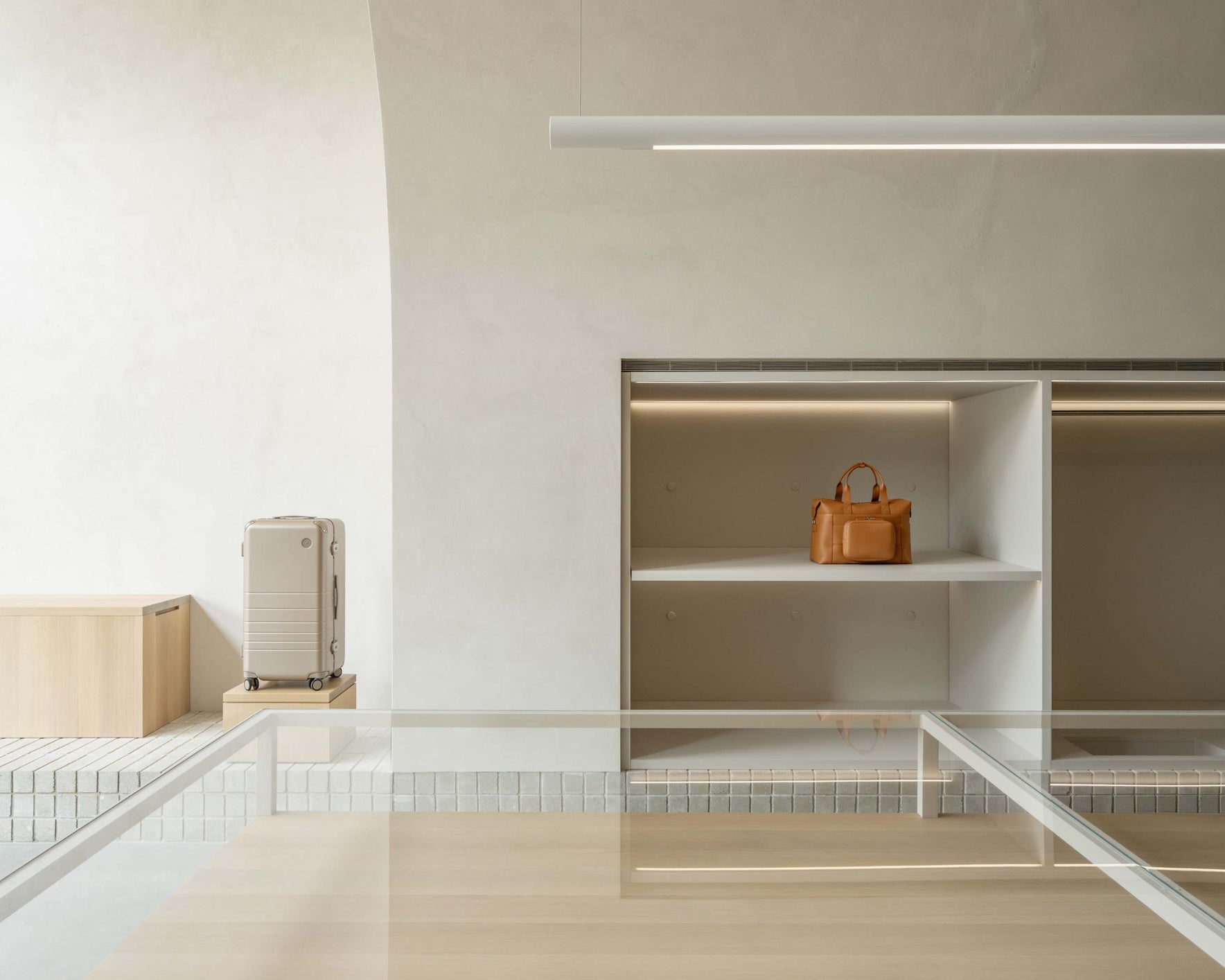 A minimalist display with a hybrid trunk in champagne on a small podium and a metro duffel in saddle tan (vegan leather) placed on an open shelf. The store has clean lines, neutral tones, and soft lighting that highlights the products.