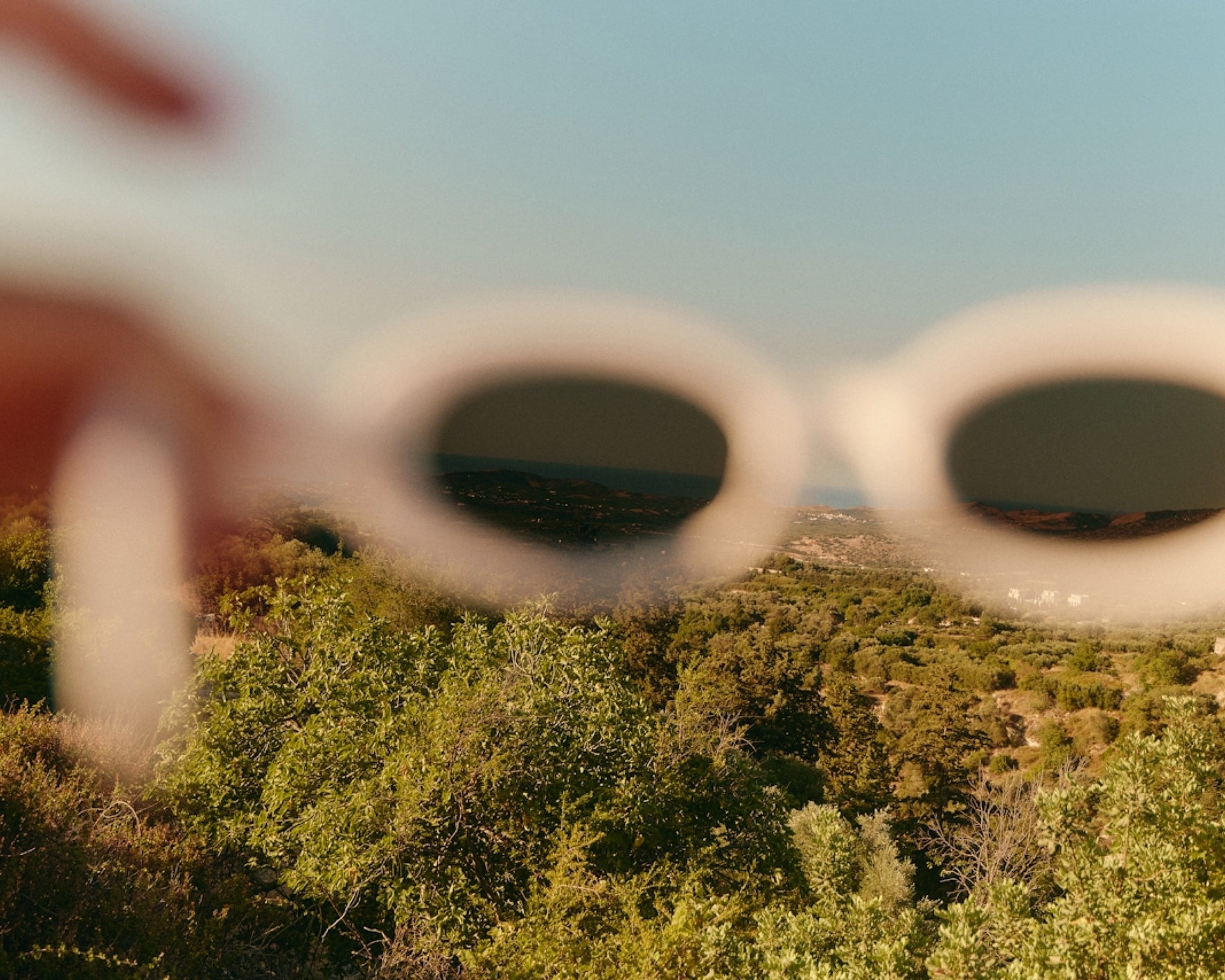 A creative perspective shot through oval-shaped white sunglasses, revealing a lush, green landscape with trees, hills, and a blue sky in the background. The edges of the sunglasses are blurred, adding depth to the scene.