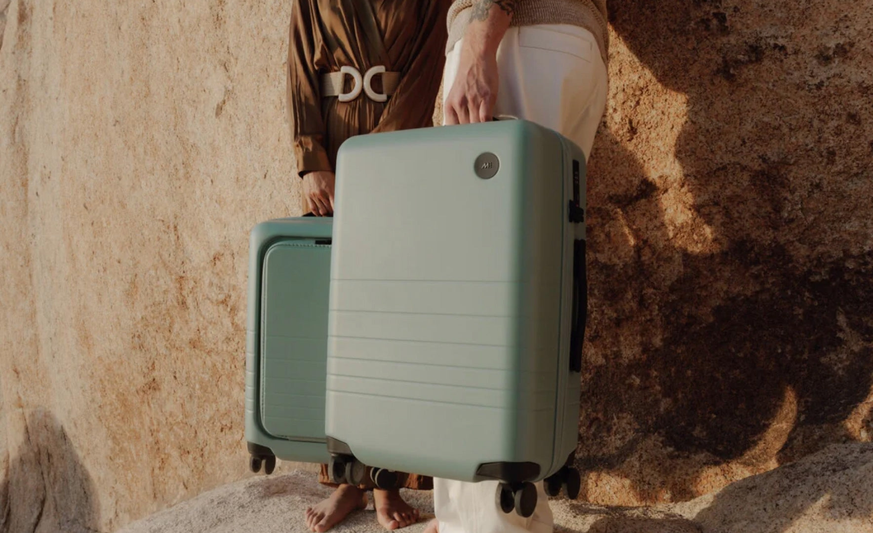 Two travellers stand by a rugged rock formation, holding a Carry-On and Carry-On Pro in Sage Green.