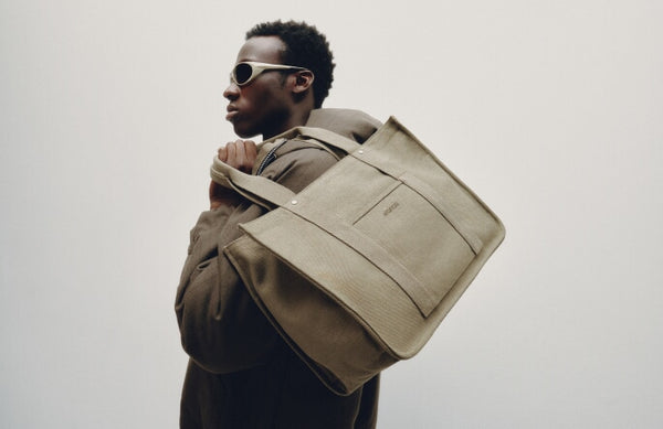A man wearing a brown jacket and sunglasses carries a Monos Gesso Tote in Moss slung over his shoulder.