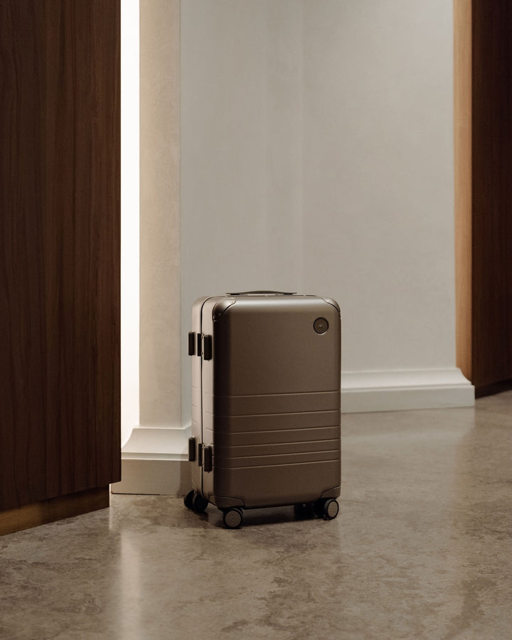 A hybrid carry-on in silver is positioned between a doorway, in a warm, indoor setting.