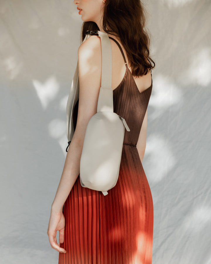 A woman wearing a flowing, pleated dress in shades of red and brown, carrying a metro sling in ivory (vegan leather) over her shoulder, with soft dappled light creating a gentle background.