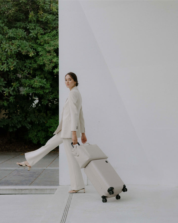 A woman in a white suit pulling a carry-on in stellar white with metro duffel in ivory (vegan leather) on top, walking along a modern architectural backdrop with green foliage in the background.