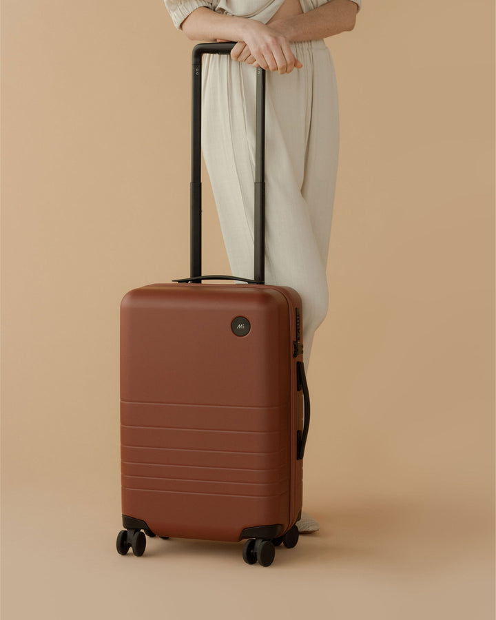 A model in beige attire stands next to a carry-on in terracotta with the telescopic handle extended, set against a warm beige background.