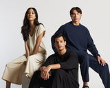 Three models seated on cubes in a studio. The woman wears a sevilla top and sevilla pants in cream, while the two men beside her are dressed in kyoto long sleeve and kyoto pants in navy and black.