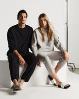 A man and woman seated side by side on white cubes in a minimalist studio, both dressed in kyoto long sleeve and kyoto pants. The man wears a kyoto long sleeve and kyoto pants in black with green sandals, while the woman is in kyoto long sleeve and kyoto pants in mist with white slides.