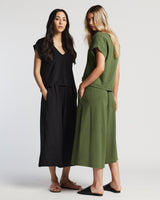 Two women stand facing away from each other in sevilla top and sevilla pants. One is dressed in all black, and the other in cypress green. They have each of their hands in their pockets, standing tall and relaxed.