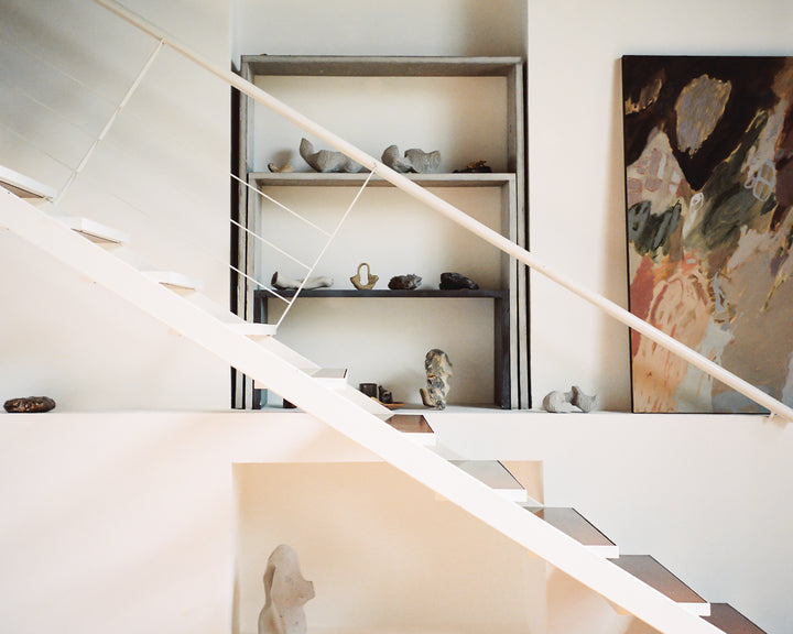 A clean, modern interior with a white staircase leading up beside a display of abstract sculptures on shelves and a colourful painting, creating a serene, artistic atmosphere.