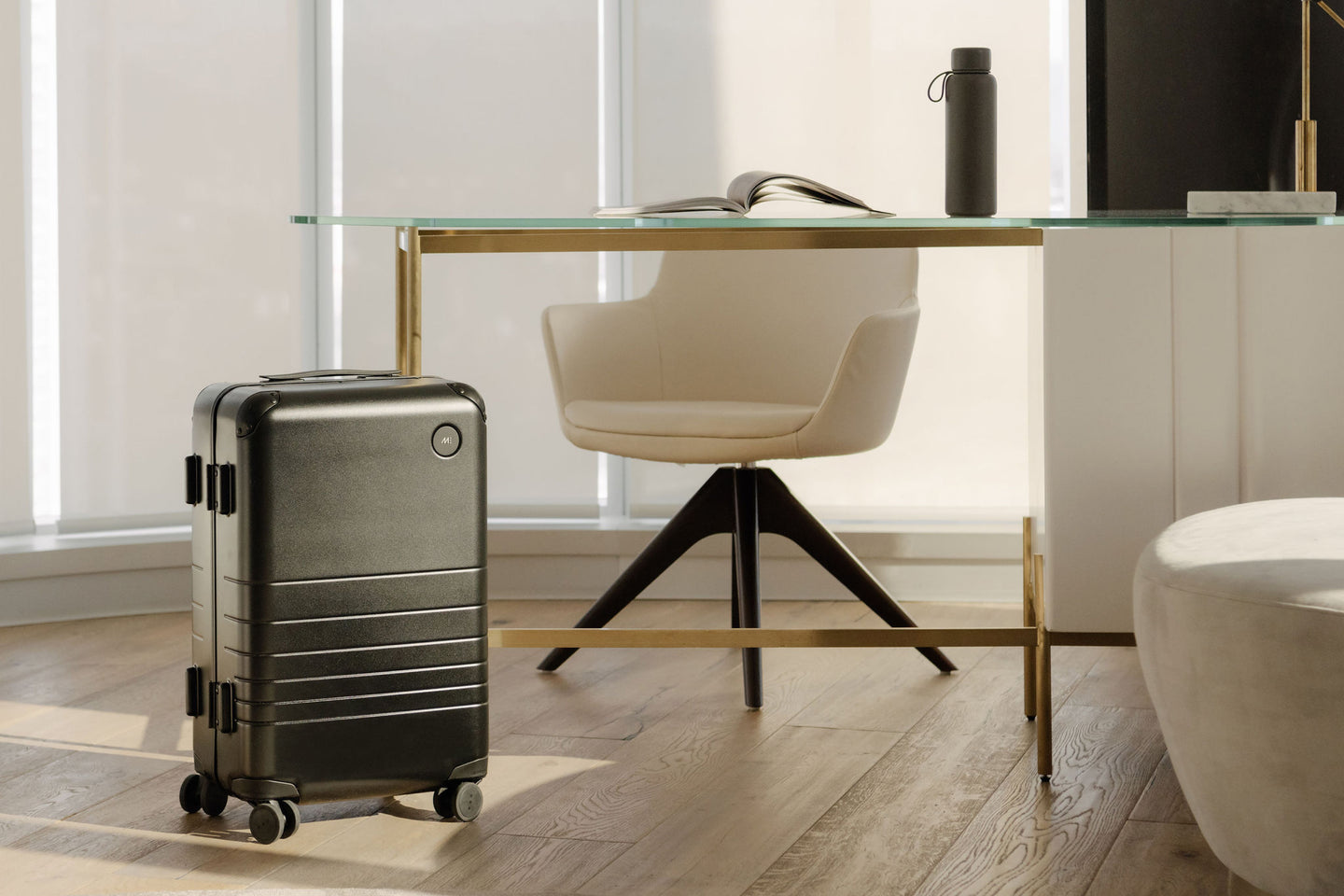 A hybrid carry-on in obsidian beside a modern glass and gold desk in a bright, stylish room.