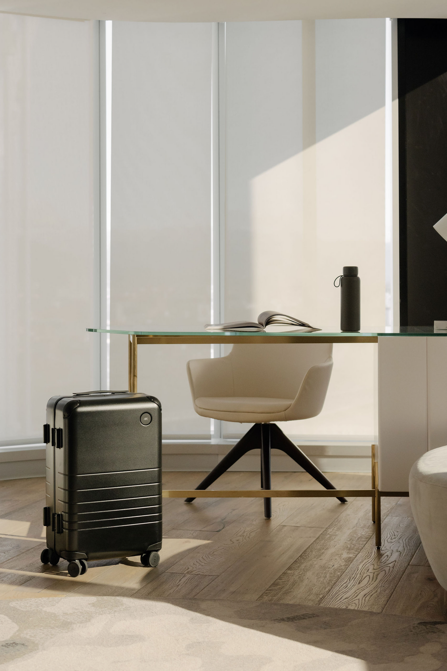 A hybrid carry-on in obsidian beside a modern glass and gold desk in a bright, stylish room.