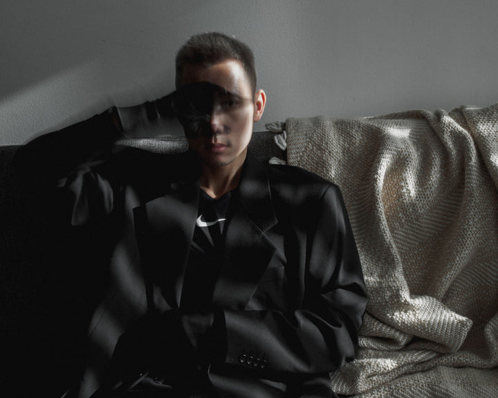 A person dressed in black sits on a sofa, partially obscured by shadows, conveying a moody and introspective vibe.