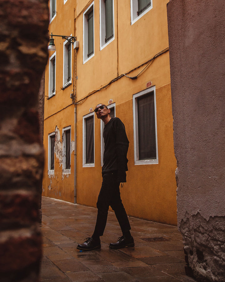 A stylish man walks down a narrow European street with orange buildings, dressed in dark attire and sunglasses, embodying a casual, urban look.