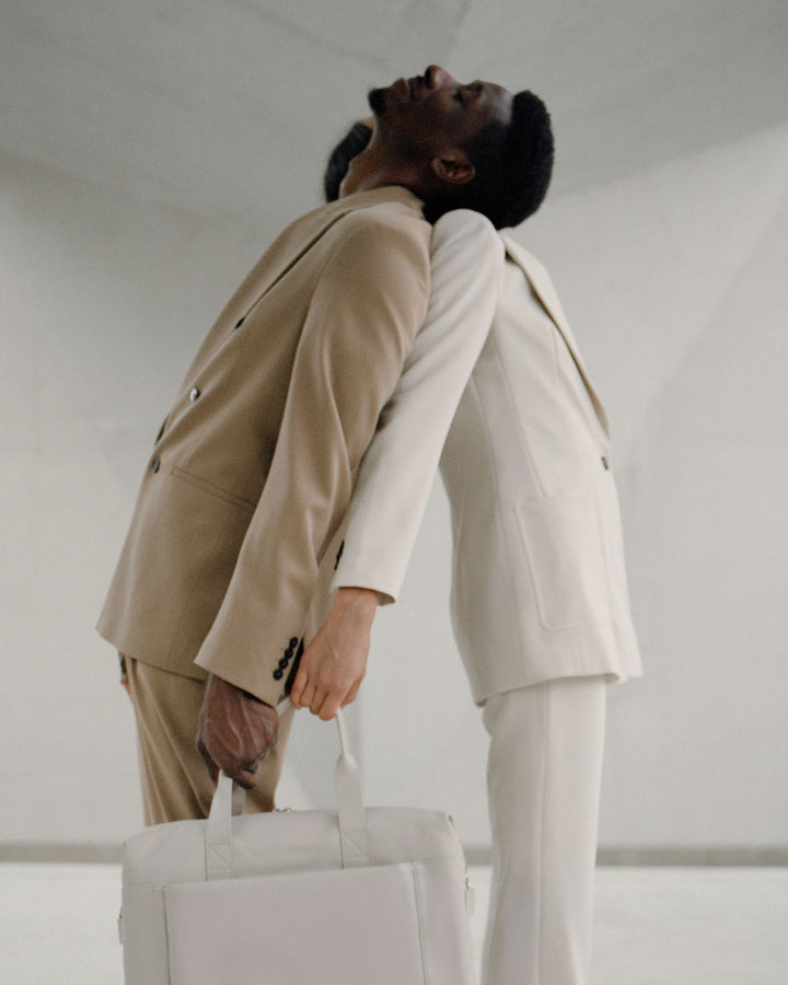 A man in a beige suit and a woman in a white suit leaning back against each other, both holding a metro duffel in ivory (vegan leather) set against a clean, white background.