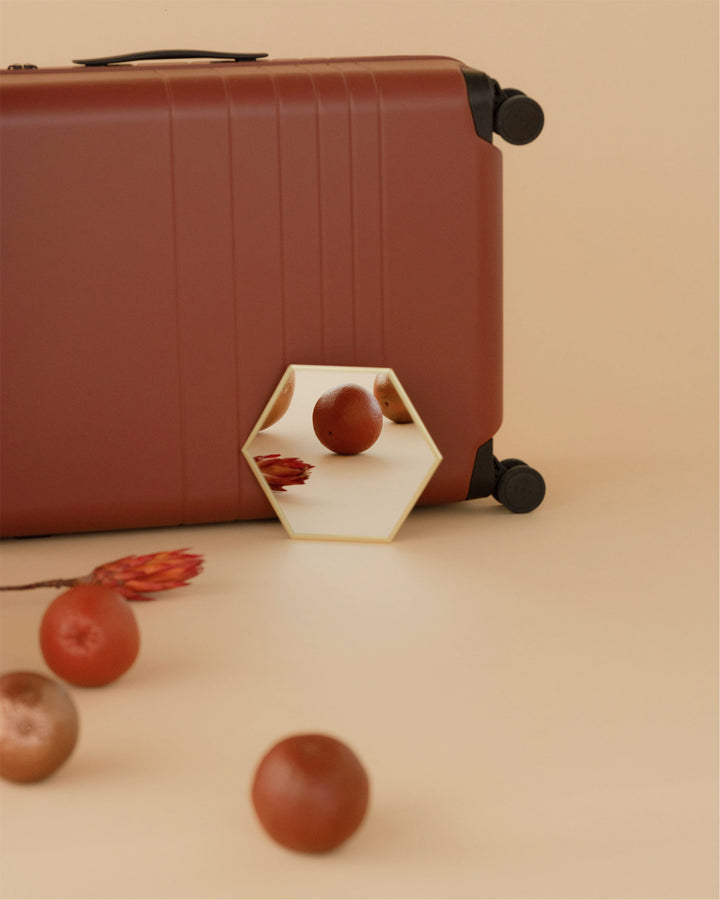 Close-up of a carry-on in terracotta with a hexagonal mirror leaning against it, reflecting a round, earthy-toned ornament alongside other decorative objects on a soft beige backdrop.