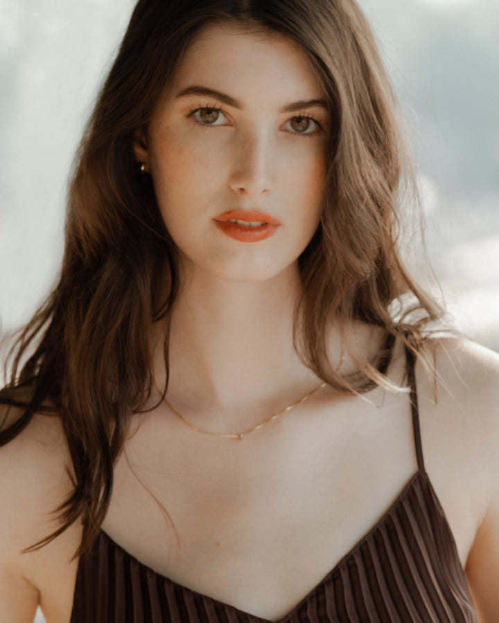 A close-up portrait of a woman with wavy brown hair and a subtle necklace, wearing a pleated dark brown top, looking directly into the camera with a calm expression.