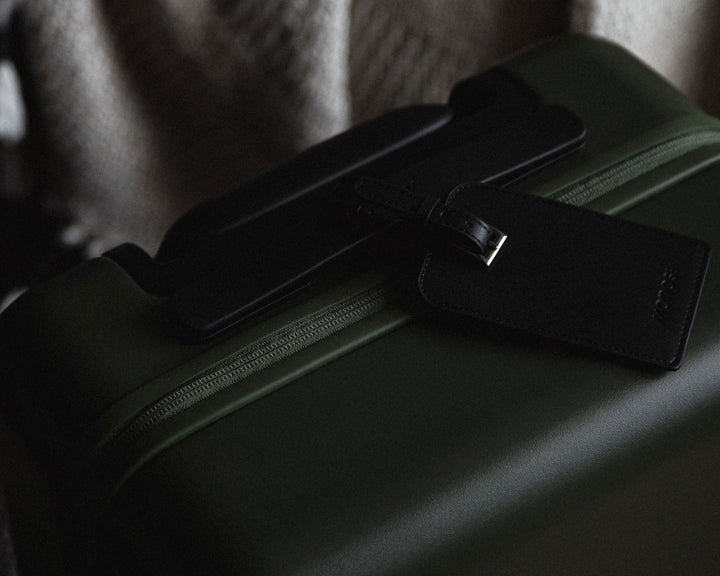 Close-up of a carry-on in olive green adorned with a black vegan leather luggage tag, placed in a cozy setting with a knitted blanket in the background.