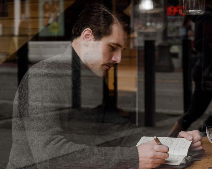 A man in a sweater and overcoat, seen through a café window, sits thoughtfully while writing in a notebook, exuding a reflective, urban vibe.