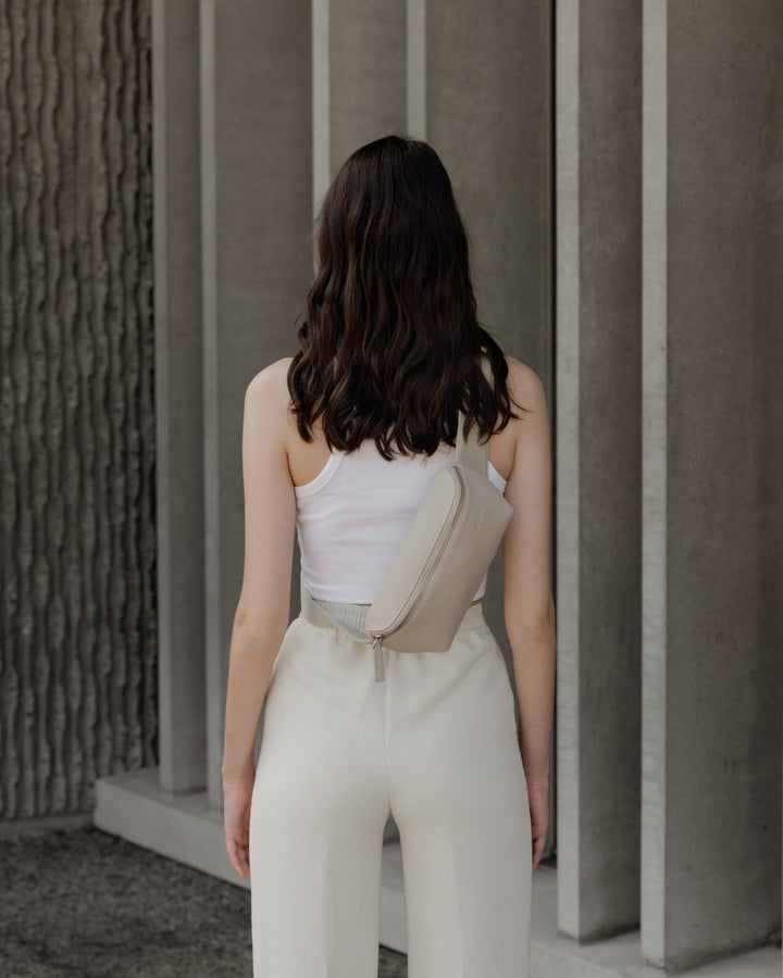 The back view of a woman in a white crop top and wide-leg pants, with a metro sling in ivory (vegan leather) strapped across her back, standing against a minimalist concrete background.