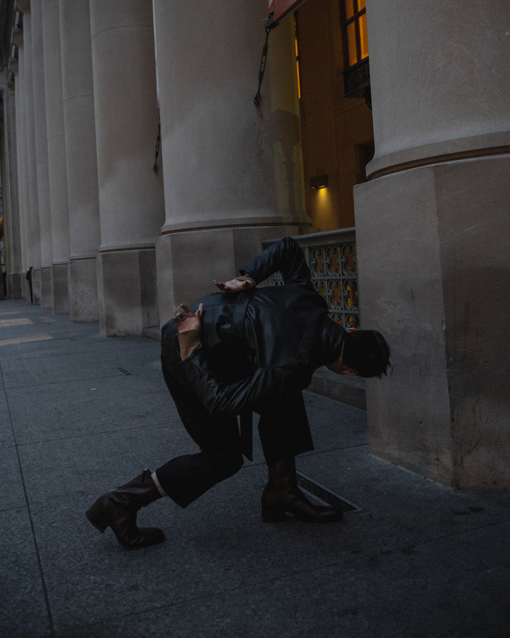 A person in a black leather outfit leans dramatically against a columned building at dusk, capturing a moment of dynamic motion in an urban environment.