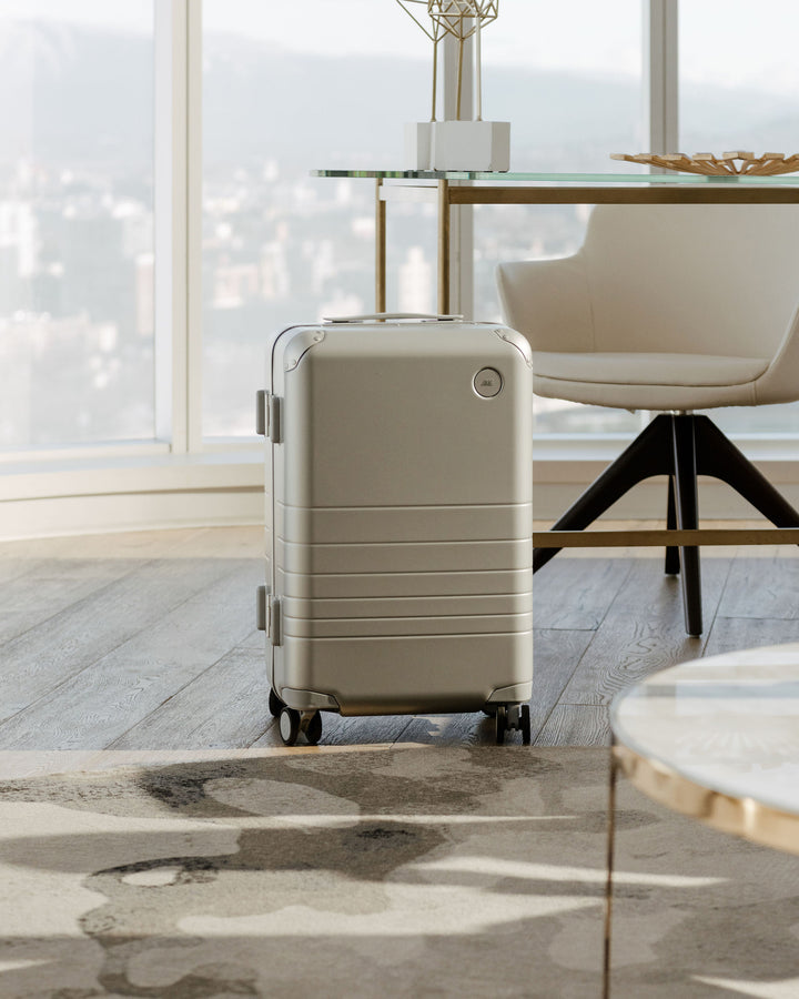 A hybrid carry-on in champagne stands on a wooden floor in a modern room with a large window, contrasting elegantly with the minimalist decor.