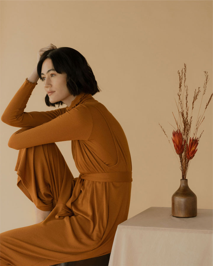 A model seated in a contemplative pose, dressed in a rust-coloured outfit, next to a table with dried floral decor, all set against a warm beige background.