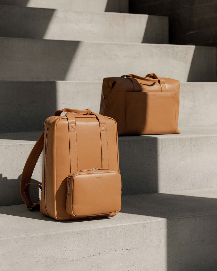 A metro backpack and metro duffel in saddle tan (vegan leather) displayed on concrete steps, highlighting their clean design and structured form.