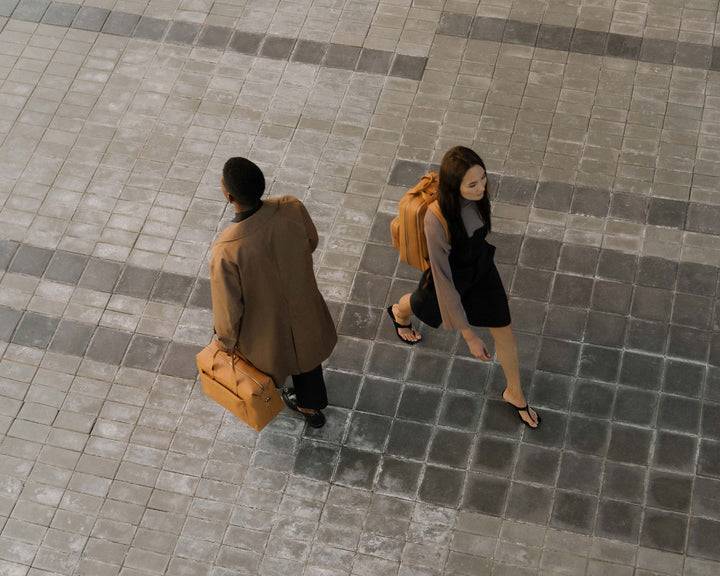 An overhead shot of a man in a brown coat and a woman in a black dress walking across a tiled courtyard, each carrying a metro duffel and metro backpack in saddle tan (vegan leather), creating a minimalist, urban aesthetic.