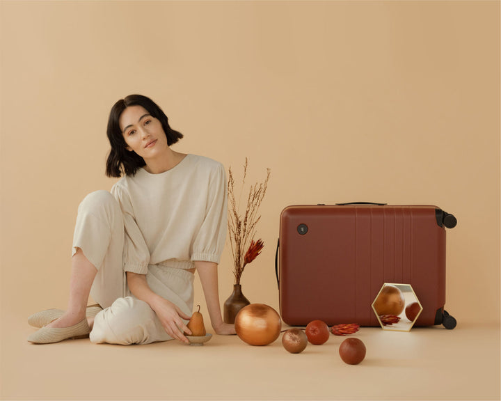 A model in beige attire sits on the floor, engaging with various decorative objects in copper and terracotta hues, alongside a check-in medium in terracotta and a small vase with dried florals.