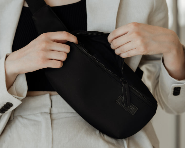 A close-up of a woman in a light blazer and black top, opening a metro sling in carbon black, highlighting the bag’s simple and functional design.