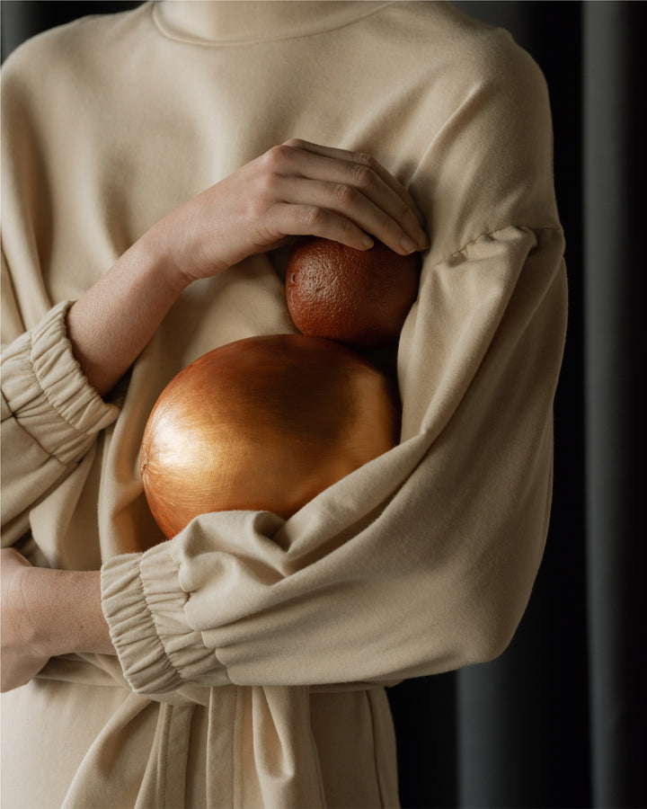 Close-up of a model’s arms cradling a large copper-coloured ornament and a smaller round decoration, against the backdrop of a beige, gathered fabric outfit.