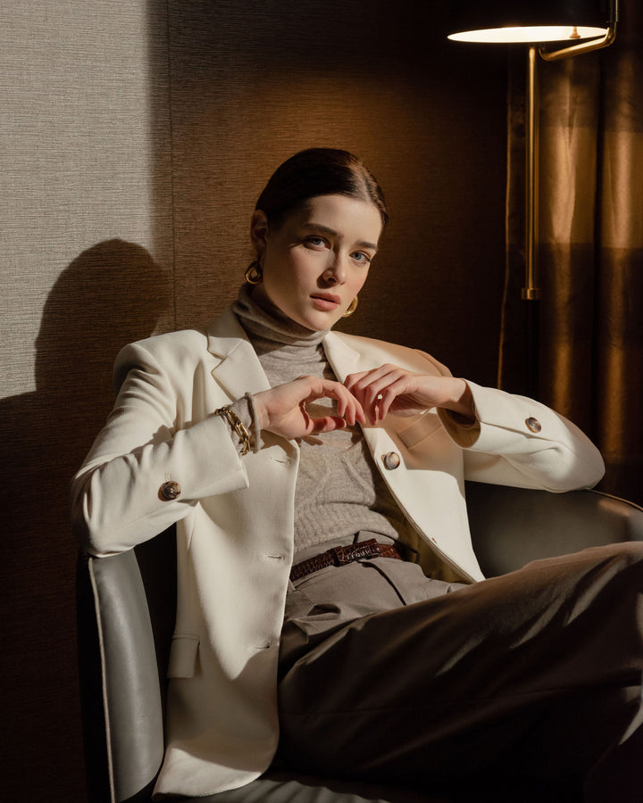  A woman in a tailored white blazer and turtleneck sits on a chair in a softly lit room, looking confidently toward the camera.