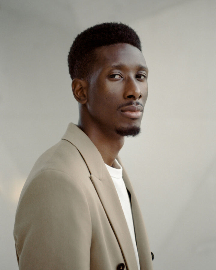 A close-up portrait of a man in a beige double-breasted blazer, looking at the camera with a serious expression, against a softly blurred background.