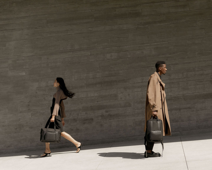 A man in a tan trench coat and a woman in a black dress walking side by side, each holding a metro duffel and metro backpack in carbon black, against a dark concrete wall.