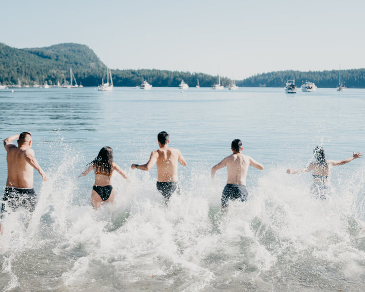 Five people, dressed in swimwear, enthusiastically run into the water, creating splashes as they embrace the joy of a sunny day by the lake.