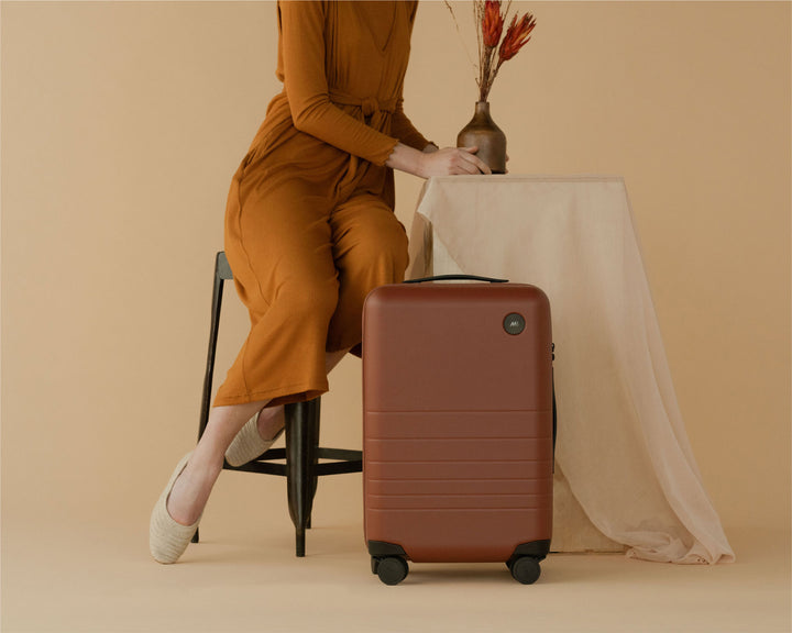 A woman wearing a rust-coloured dress sits on a chair beside a carry-on in terracotta, with her feet crossed and resting gracefully. A small table with a vase holding dried flowers adds an elegant, earthy touch to the scene.