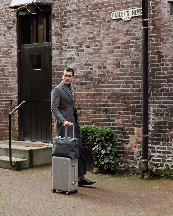 A man in a grey coat pulls a hybrid carry-on in silver along a brick alley, blending sophisticated travel style with an urban setting.