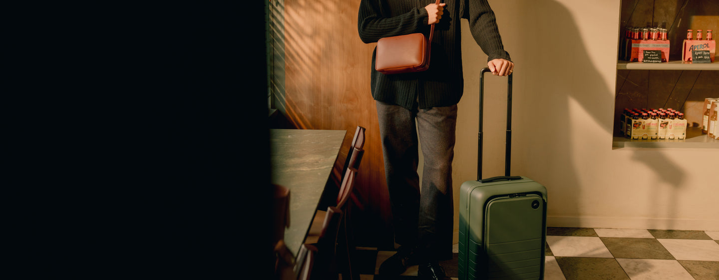 A traveler dressed in dark tones stands in a sunlit café, holding the Monos Carry-On Pro in olive green.