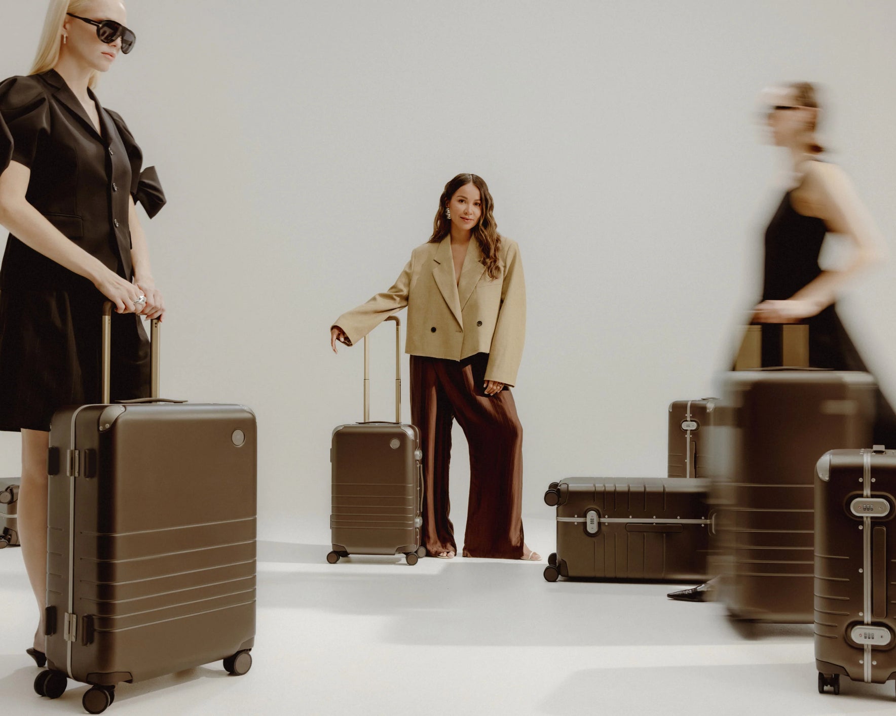 Three women, each with Hybrid luggage in Adobe Brown, are posed. Two are in motion, creating a dynamic blur, while one stands still.