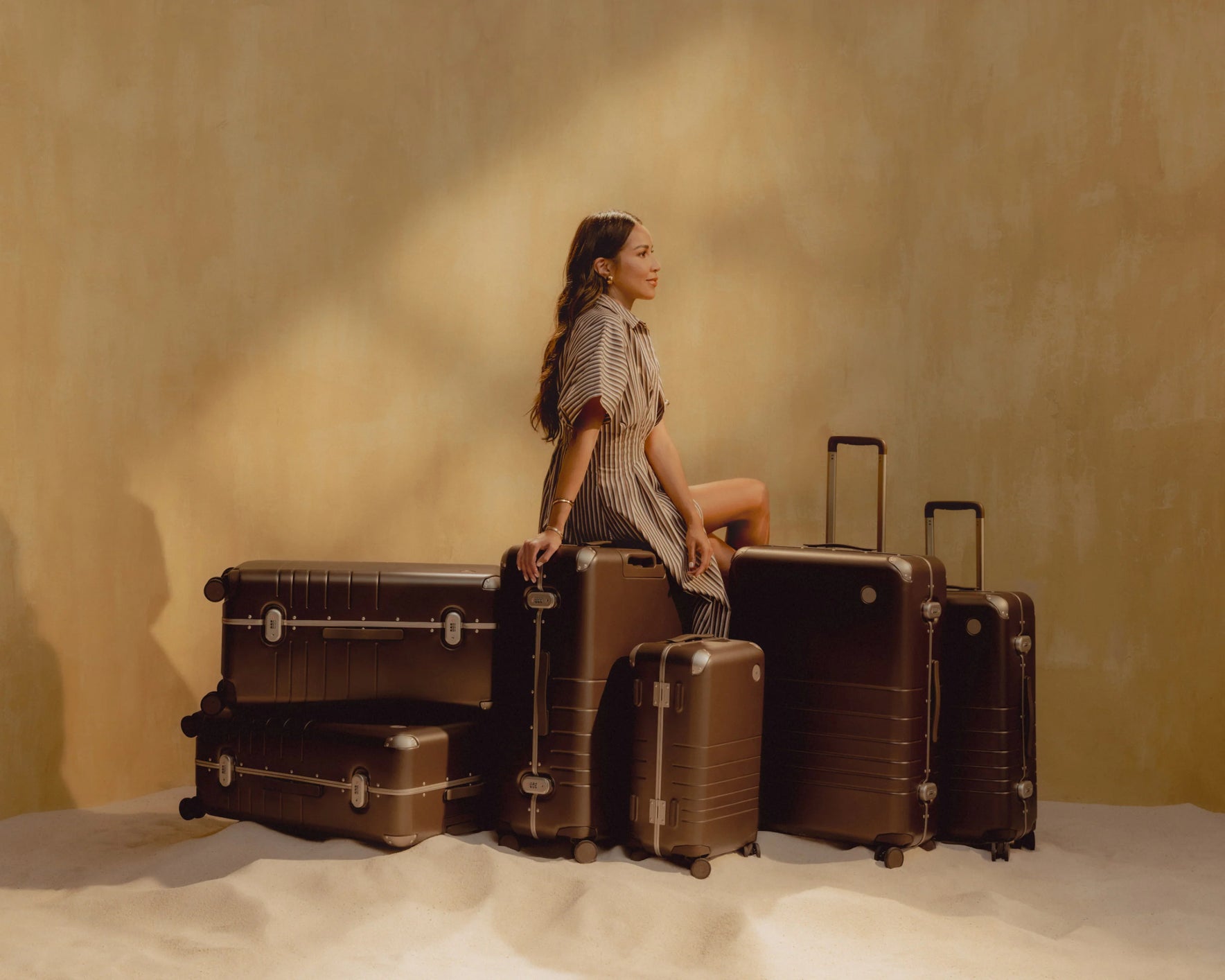 A woman sits on a stack of Hybrid luggage in Adobe Brown, surrounded by more Hybrid luggage in Adobe Brown, in a warm, sandy setting.