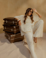 A woman wearing a white outfit, sits in a chair with her hand shielding her eyes from the sun. Three Hybrid suitcases in Adobe Brown are stacked beside her against a sandy-coloured backdrop.