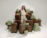 A woman stands among stacked Hybrid suitcases in Adobe Brown and bags and accessories in Cactus.