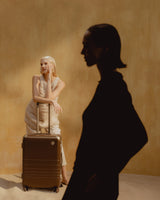 A woman leans on a Hybrid Check-In Medium in Adobe Brown in front of a sandy-coloured backdrop. A second woman is silhouetted in the foreground.