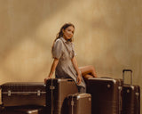 A woman sits on a stack of Hybrid luggage in Adobe Brown, surrounded by more Hybrid luggage in Adobe Brown, in a warm setting.