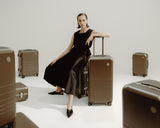 A woman dressed in black sits on a Hybrid luggage in Adobe Brown, surrounded by more various Hybrid luggage in Adobe Brown.