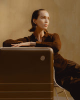 A woman leans on a Hybrid Check-In Large in Adobe Brown while sitting on a chair against a warm, sandy-toned background.