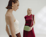 Two women are standing against a white background. One in the foreground looking down. The woman in the background carries a Metro Crossbody in Cactus (Vegan Leather).