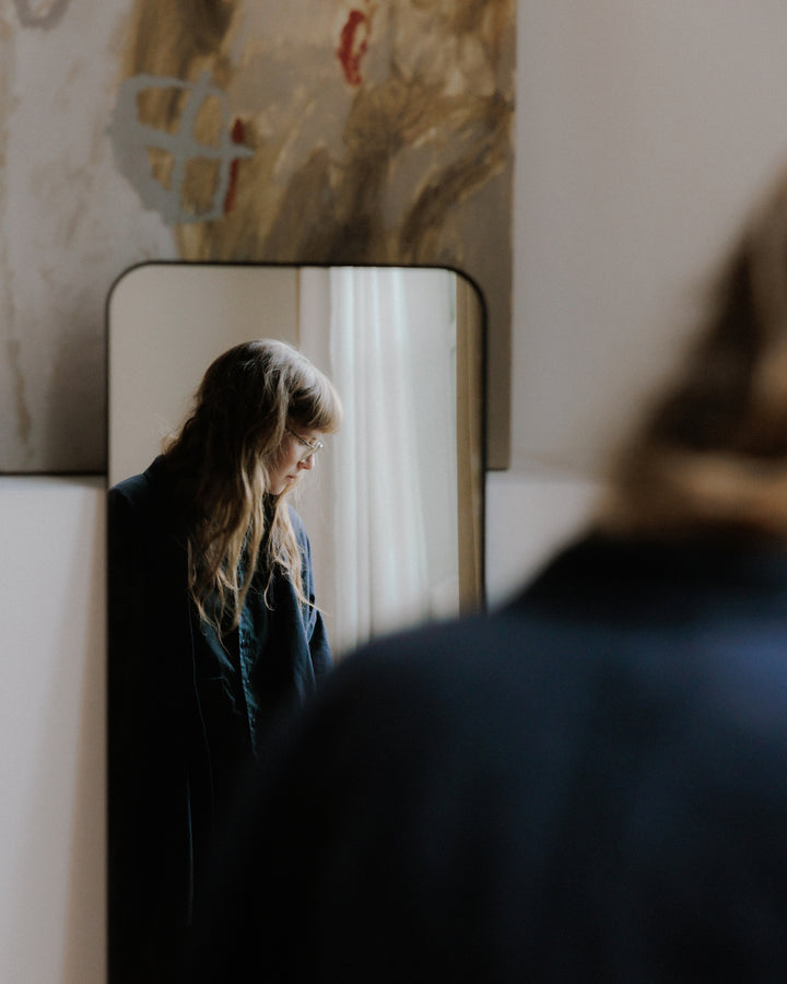 A woman is reflected in a mirror, her expression soft as she looks down, with abstract artwork visible behind her, evoking a quiet, contemplative mood.