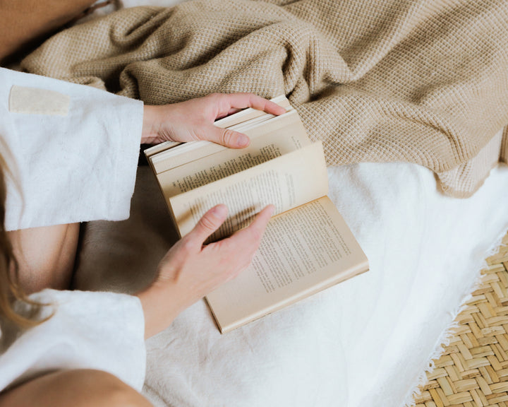 A close-up of hands holding an open book, with a warm blanket in the background, creating a sense of comfort and relaxation.